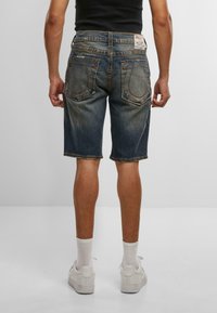 Muž oblečený vo vypasovaných modrých džínsových kraťasoch, bielych ponožkách a bielych teniskách, zobrazený zozadu na svetlom pozadí.