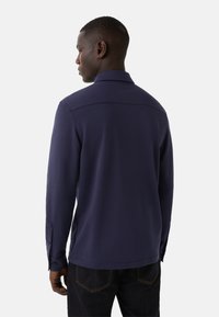 Man die met zijn rug naar de camera staat, in een marineblauwe overhemd met lange mouwen en kraag, en donkere jeans, tegen een effen lichte achtergrond.