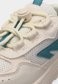 Detaljan prikaz bijele tenisice od mrežastog materijala i antilopa s tirkiznim naglascima, elastičnim vezicama i bež kopčom na vrhu.