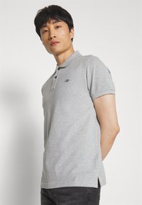 Man wearing light grey short-sleeve polo shirt with small logo, standing against plain white background, looking slightly upward.