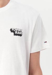 White cotton t-shirt featuring a black "TOMMY JEANS" logo on the left chest. Short sleeves with a small three-striped accent on the sleeve.