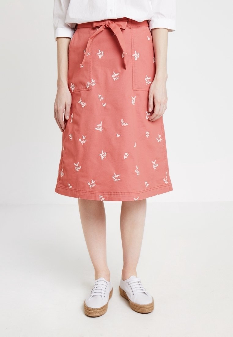 Jupe corail longueur genoux avec imprimés floraux blancs, poches avant et taille nouée, portée avec une chemise blanche et des baskets à plateforme blanches.