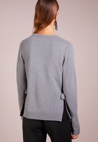 Grå stickad tröja med långa ärmar, rund halsringning, sidofrack och en strukturerad yta. Har ett lager av svart underplagg som syns vid fållen.