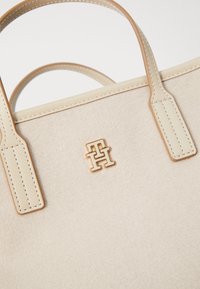 Bolsa de camurça bege com acabamentos em tom de ouro. Apresenta duas alças robustas e um destaque com logotipo. Textura suave com detalhes cosidos.