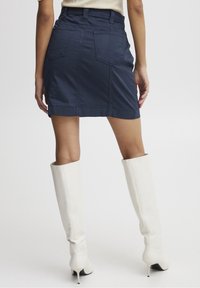 Jupe mini en denim marine avec un design ajusté, dotée de deux poches arrière et de coutures visibles, associée à des bottes blanches à talons jusqu'au genou.