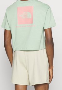 Miętowo-zielony, krótki t-shirt wykonany z miękkiej tkaniny, z różowym graficznym prostokątem, na którym znajduje się napis "THE NORTH FACE" oraz "NEVER STOP EXPLORING" poniżej.