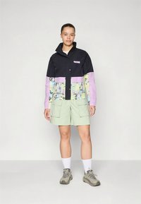 Kleurrijk jack met zwarte bovenkant, pastelpaarse mouwen met bloemenpatroon, gecombineerd met lichtgroene cargoshorts en grijze schoenen.