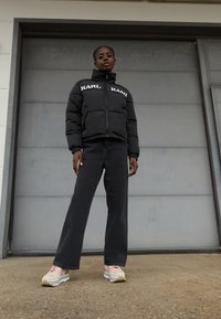 Giubbotto imbottito nero con logo bianco, colletto alto; jeans scuri; sneakers rosa chiaro con suola bianca, design a strati sui lati.