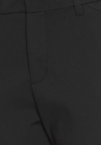 Détail d'un pantalon noir sur mesure montrant la couture avant, la ceinture et la passants sur un tissu lisse.