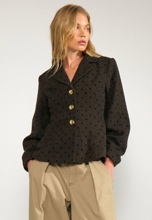 Femme portant une veste marron foncé à pois avec trois boutons et un pantalon beige taille haute, les mains dans les poches, regardant sur le côté.