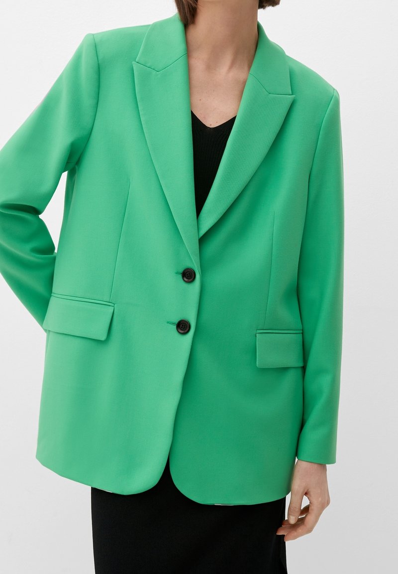 Personne portant un blazer vert vif avec des boutons noirs par-dessus un haut noir, main posée sur la hanche, sur un fond uni.
