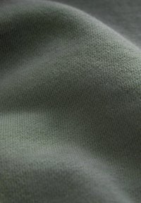 Zbliżenie na gładką, oliwkowozieloną dzianinę z drobnymi, równymi ściegami i delikatnymi fałdami tworzącymi miękkie cienie.