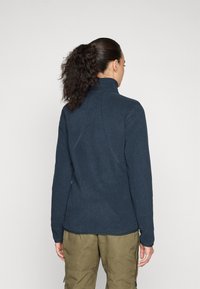 Dark blue fleece jacket with a high collar, textured finish, and fitted shape. Features flat seams and a straight hem. Paired with cargo pants.
