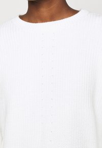 Close-up van een persoon die een witte ribgebreide trui draagt met een ronde hals en subtiele verticale stikdetails.