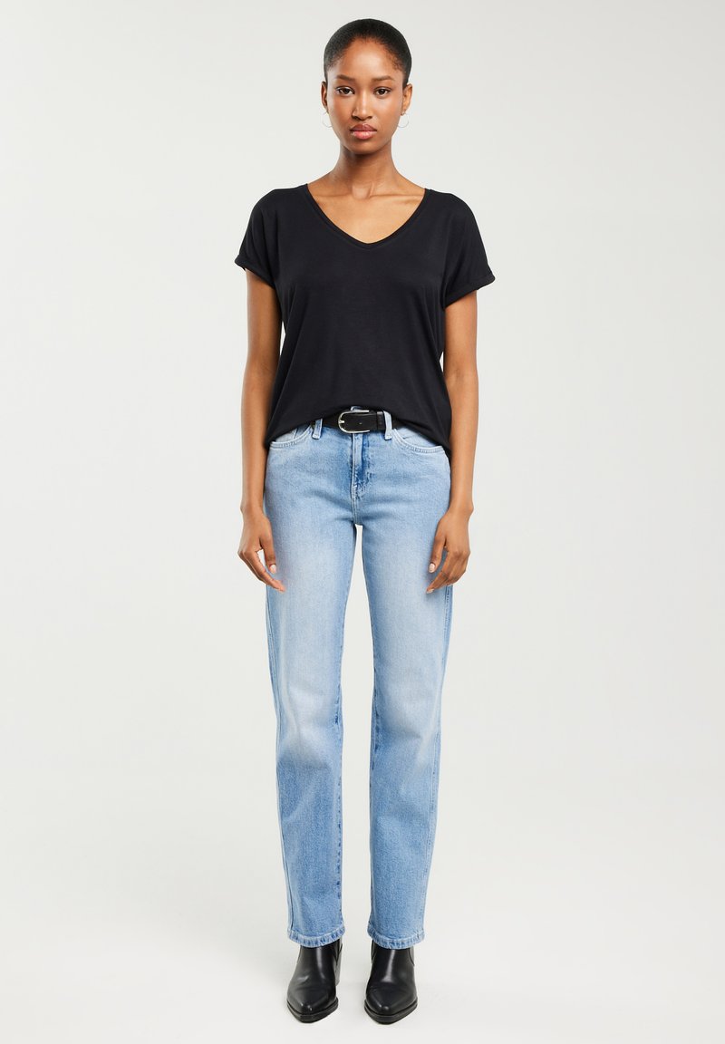 Sieviete, tērpusies melnā T-kreklā ar V veida kakla izgriezumu, gaiši zilās džinsās ar melnu jostu un melnās potīšu zābakos, stāvot pret vienveidīgu gaišu fonu.