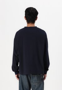 Μακρυμάνικη ναυτική μπλούζα, κατασκευασμένη από βαμβάκι, άνετη εφαρμογή, στρογγυλή λαιμόκοψη, χαμηλωμένοι ώμοι, πίσω όψη, συνδυασμένη με μπλε τζιν παντελόνι.