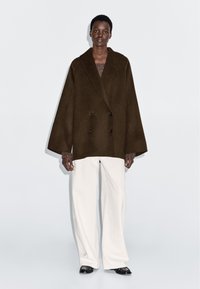 Manteau en laine brun avec un design croisé, de grands revers et des manches larges, associé à un pantalon blanc à jambes larges et des chaussures noires.