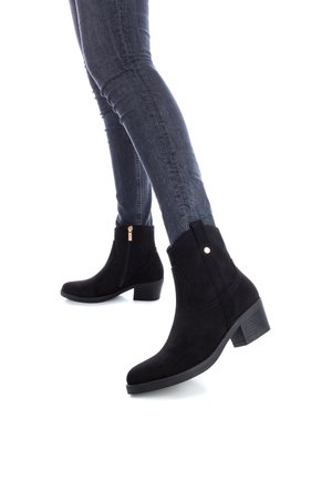 Botines de ante negros con tacón bajo en bloque, cremallera lateral y detalles de costura sutiles. Llevados con jeans ajustados oscuros.