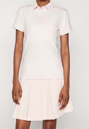 Vrouw draagt een lichtroze polo met korte mouwen en een bijpassende geplooide rok, staand tegen een effen witte achtergrond.