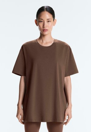 Žena s hladkými vlasmi, ktorá nosí veľké hnedé tričko s krátkym rukávom, stojaca pred jednoduchým svetlým pozadím.