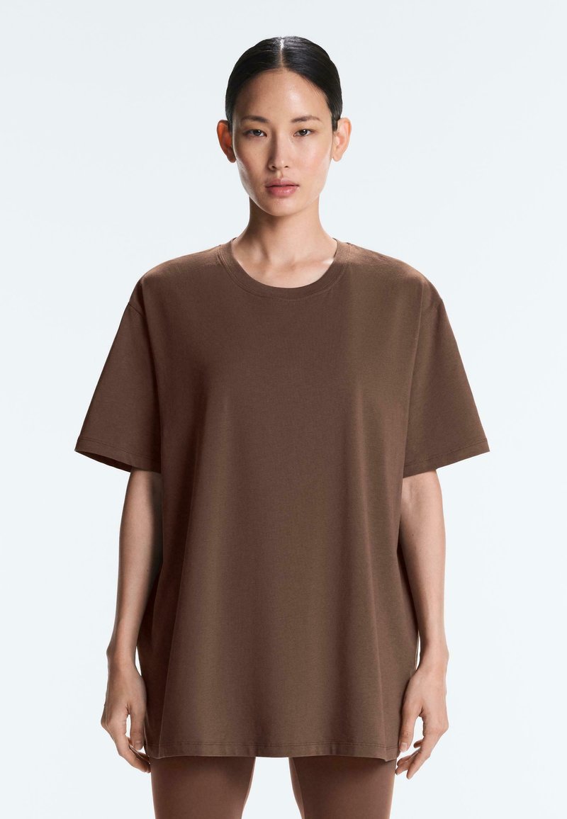 Femme aux cheveux lisses portant un t-shirt marron à manches courtes oversize, debout devant un fond clair uni.