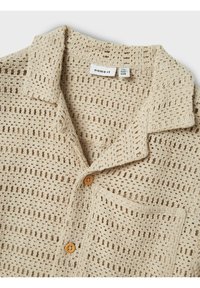 Chemise en tricot beige avec un tissage aéré, col court, une poche poitrine et des boutons marron. Tissu texturé avec un motif uniforme.