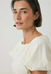 Blusa bianca e strutturata con scollatura tonda e maniche a palloncino, indossata da una modella con capelli corti e scuri, su uno sfondo neutro.