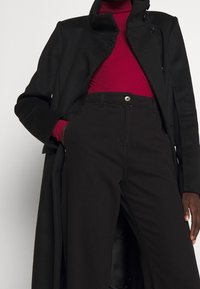 Cappotto nero con colletto alto, dotato di doppi bottoni decorativi, indossato sopra un maglione rosso a collo alto e pantaloni neri. Tessuto liscio e vestibilità su misura.