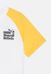 Camiseta blanca con mangas raglán amarillas, que presenta un logo gráfico negro "PUMA Board Rebels" en la parte delantera. Material de algodón suave, corte casual.