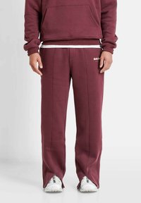 Bordeauxfarbene Jogginghose mit lockerer Passform, seitlichen Nähten, einem Logo auf dem linken Oberschenkel und Schlitzsäumen an den Knöcheln. Glatte Textur.