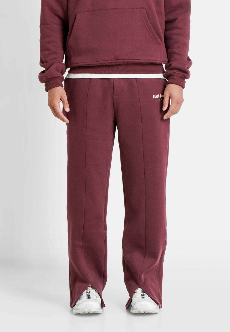 Bordeauxfarbene Jogginghose mit lockerer Passform, seitlichen Nähten, einem Logo auf dem linken Oberschenkel und Schlitzsäumen an den Knöcheln. Glatte Textur.