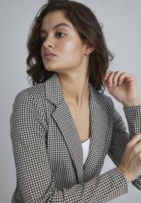 Mustvalge houndstooth mustriga bleiser, millel on krae ja sobiv disain. Tekstureeritud kangas, ühe nööbi sulgemine, koos valge särkiga all.
