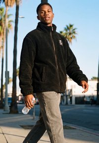 Mladý muž kráčí venku po slunečné ulici, má na sobě černou fleecovou bundu a šedé kalhoty, v pravé ruce drží láhev s vodou.