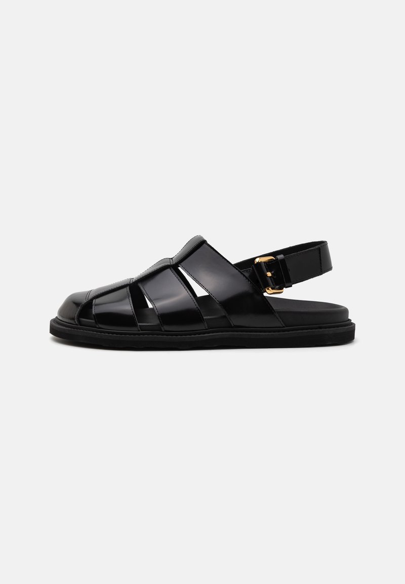 Black sandals made of glossy material featuring a strappy design, secured with a gold buckle, and a flat, textured sole.