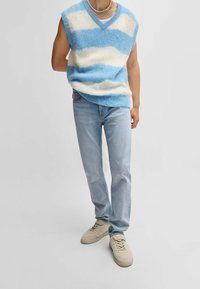 Maglione senza maniche a righe azzurro chiaro e bianco con scollo a V, abbinato a jeans in denim azzurro chiaro e sneakers beige.