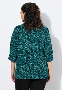 Blusa turchese con stampa leopardata, maniche a tre quarti, tessuto morbido, vestibilità rilassata e orlo arrotondato, indossata sopra pantaloni neri.