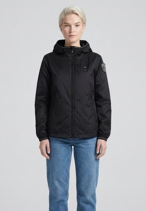 Giovane con capelli biondi corti che indossa una giacca nera con cappuccio e jeans blu, in piedi contro uno sfondo chiaro e uniforme.