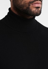 Svart stickad tröja med hög hals i ribbat material, med en slät yta och en figurnära passform.