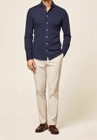 Homme portant une chemise bleu marine à boutons, un pantalon beige et des mocassins en cuir marron, debout avec une main dans la poche sur un fond uni.
