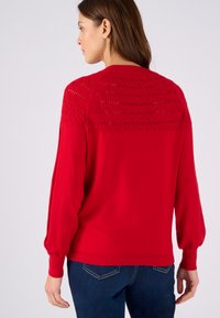 Maglione rosso lavorato a maglia con un motivo a rilievo sul giromanica, maniche a palloncino e polsini e orlo a costine; abbinato a jeans in denim blu scuro.
