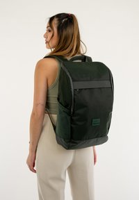 Sac à dos vert foncé au design épuré, fabriqué en tissu résistant. Il dispose d'une poignée supérieure, de bretelles réglables et d'une poche zippée à l'avant.