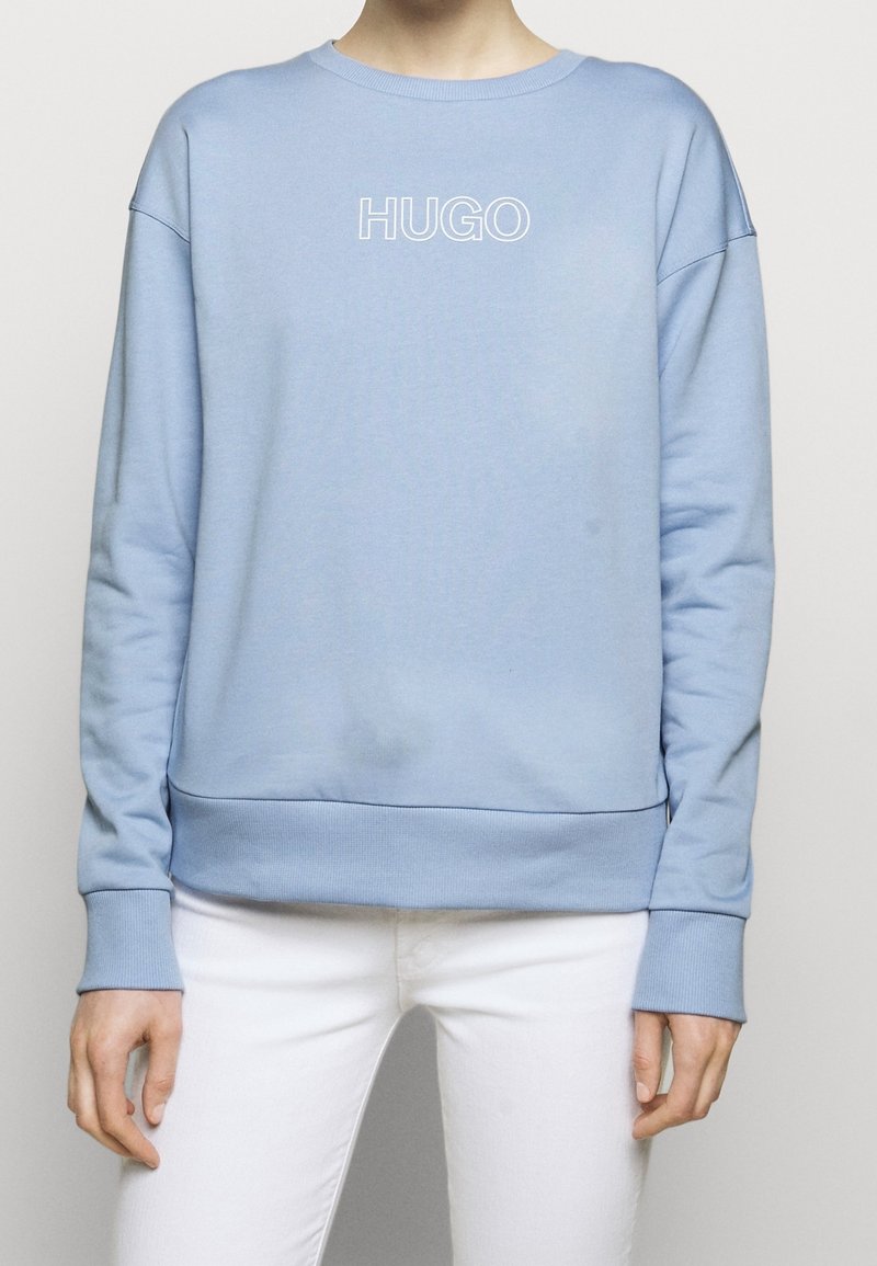 Personne portant un sweat-shirt bleu clair avec le texte "HUGO" sur le devant, assorti à un pantalon blanc, debout devant un fond uni.