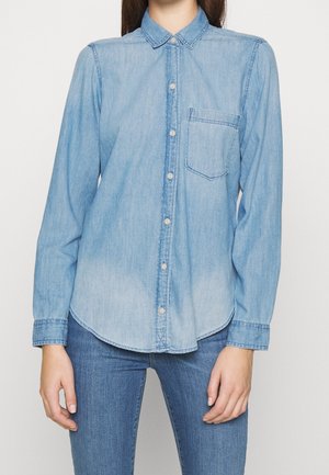 Personne portant une chemise en denim bleu clair à boutons avec une poche poitrine et un jean bleu moyen sur un fond uni.