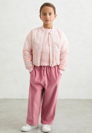 Bērns stāv ar rokām kabatās, valkājot gaiši rozā rakstainu izšūtu jaku, rozā džemperi, plašām kājām rozā bikses un baltus sporta apavus.