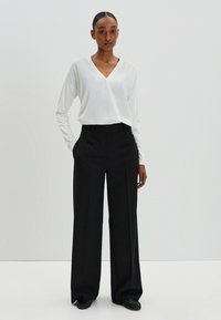 Blouse blanche à manches longues avec un col en V, associée à un pantalon noir taille haute à jambes larges, portée avec des chaussures noires à enfiler.