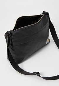 Borsa a tracolla nera realizzata in tessuto resistente, con apertura superiore con zip, tracolla regolabile e una piccola etichetta con logo sul lato.