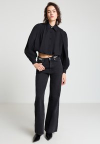 Schwarze, kurze Bluse mit Puffärmeln und Knopfleiste, kombiniert mit hoch taillierten, schwarzen Schlagjeans. Schwarze spitze Schuhe runden den Look ab.