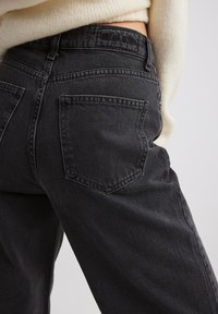 Vaqueros de mezclilla negros de cintura alta, con clásicos bolsillos traseros y un acabado texturizado. La cinturilla tiene un branding sutil.