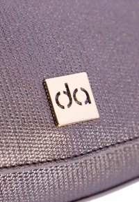 Tela texturizada gris con un sutil brillo, con una placa cuadrada de logo dorado con las letras "da" en relieve. Detalle de costura ordenada visible.