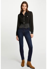 Blusa negra de manga larga con pliegues, dos bolsillos en el pecho y un dobladillo curvado. Combinada con jeans skinny de color azul oscuro y botas de tobillo marrones.